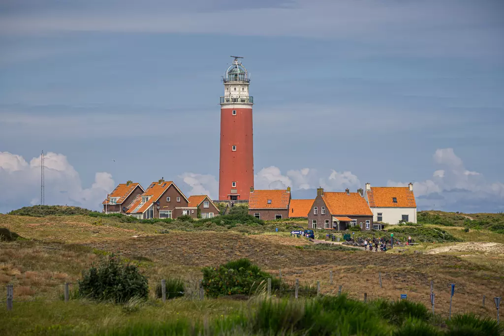 Ein rot-weißer Leuchtturm steht vor einem kleinen Dorf mit orangefarbenen Dächern, umgeben von grünen Dünen und blauen Himmel.