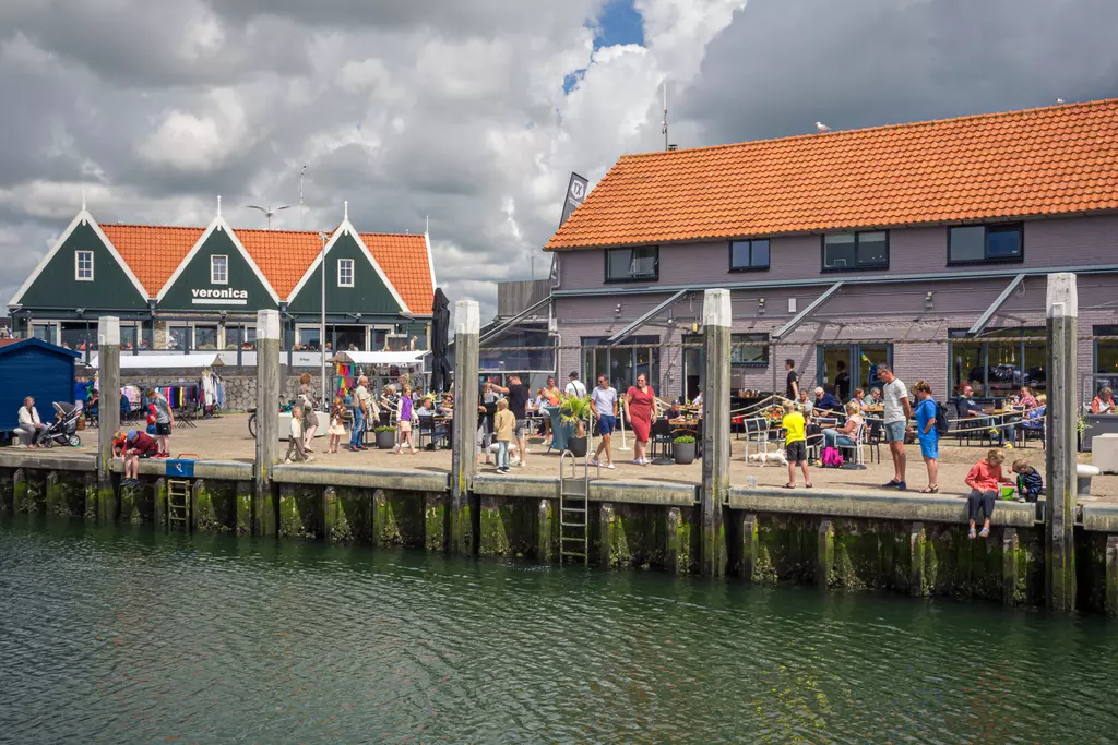 Texel: Hafen mit mehreren Menschen, die an der Uferpromenade stehen, umgeben von Gebäuden und dem Wasser im Vordergrund.