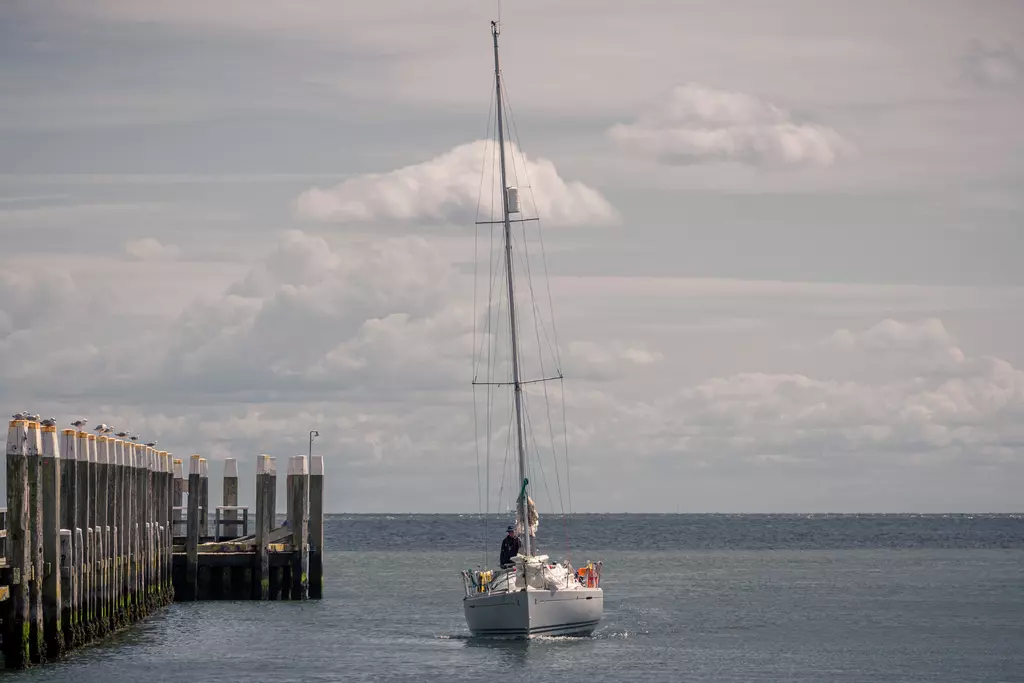 Segelboot in Texel: Ein Segelboot bewegt sich sanft im Wasser, während es sich einem Steg mit Pfählen nähert.