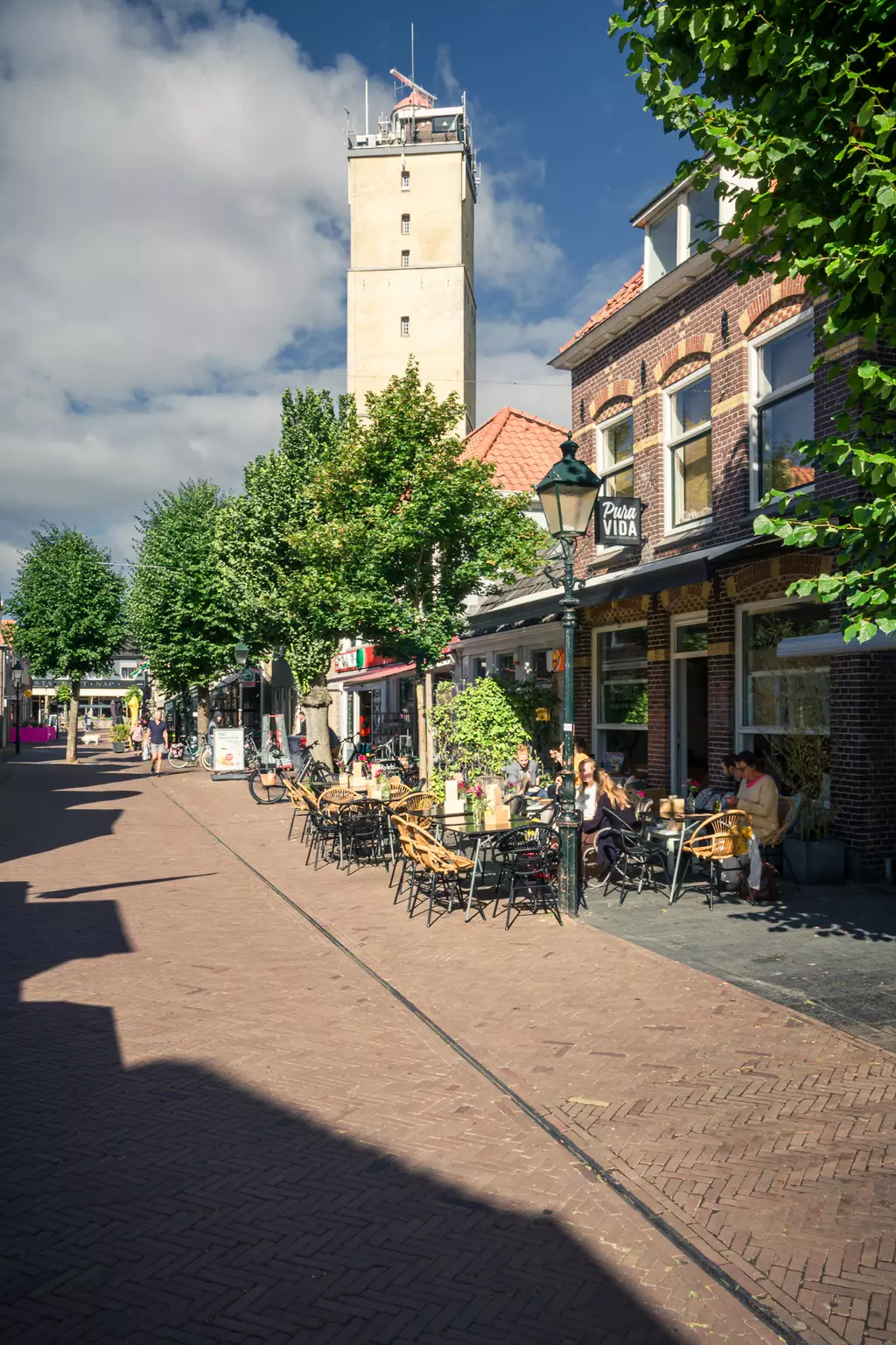 Stadtansicht auf Terschelling: Eine belebte Straße mit Cafés, Bäumen und einem hohen Gebäude im Hintergrund.