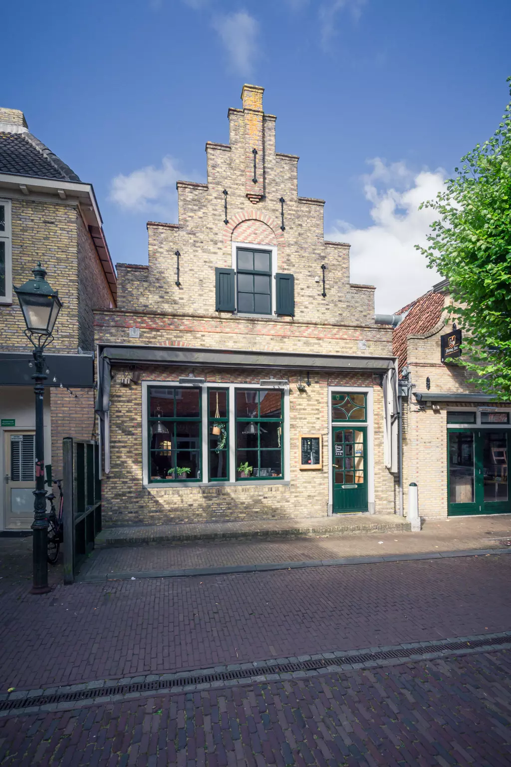 Gebäude in Terschelling: Ein historisches Backsteinhaus mit Giebeldach, großen Fenstern und grünen Türen, umgeben von gepflasterter Straße.