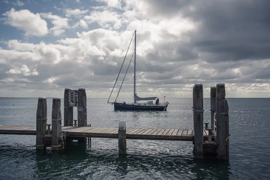 Steg auf Terschelling mit einem Segelboot im Hintergrund auf ruhigem Wasser und bewölktem Himmel.