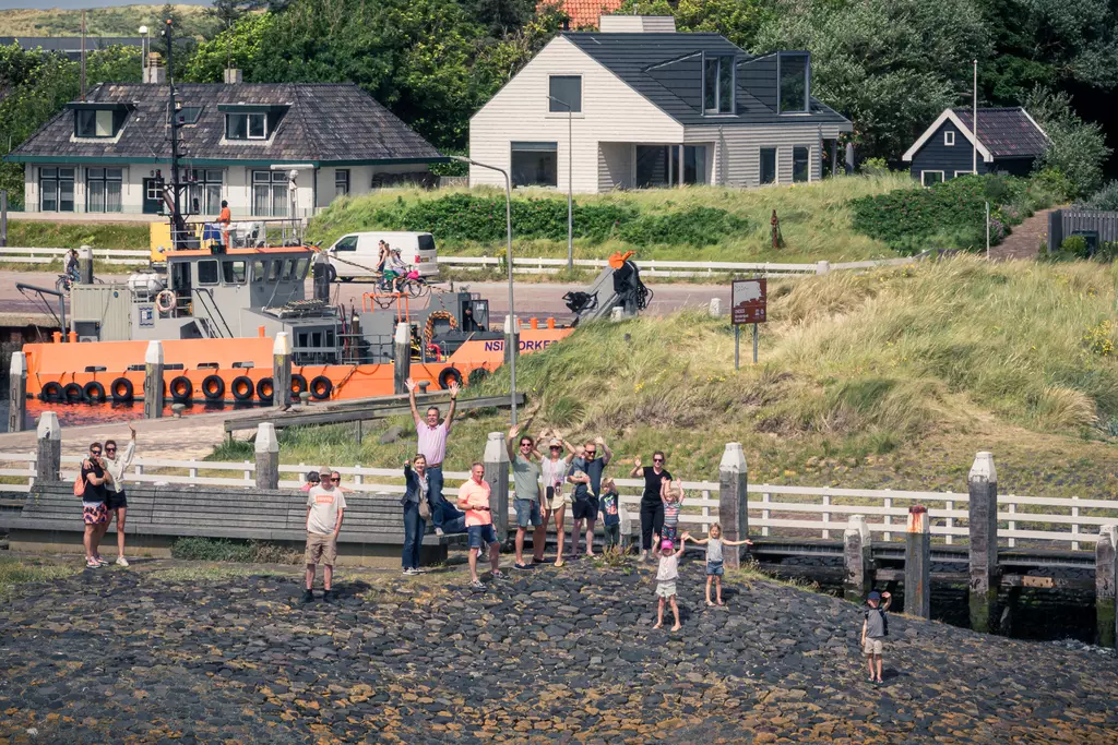 A group of people stands at the harbor, some waving. In the background, there are houses and a boat.