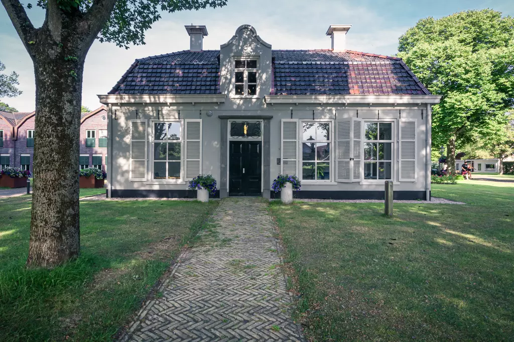 Ein weißes Haus mit einem roten Ziegeldach und großen Fenstern. Vor dem Haus führt ein gepflasterter Weg durch einen grünen Garten.