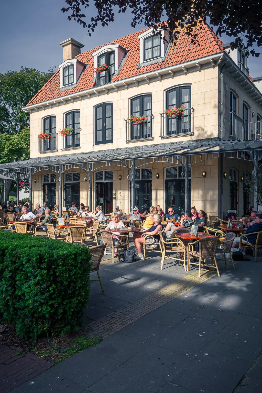 Eine große Terrasse eines Restaurants ist voller Gäste, die an Tischen sitzen und essen oder trinken, umgeben von Pflanzen.