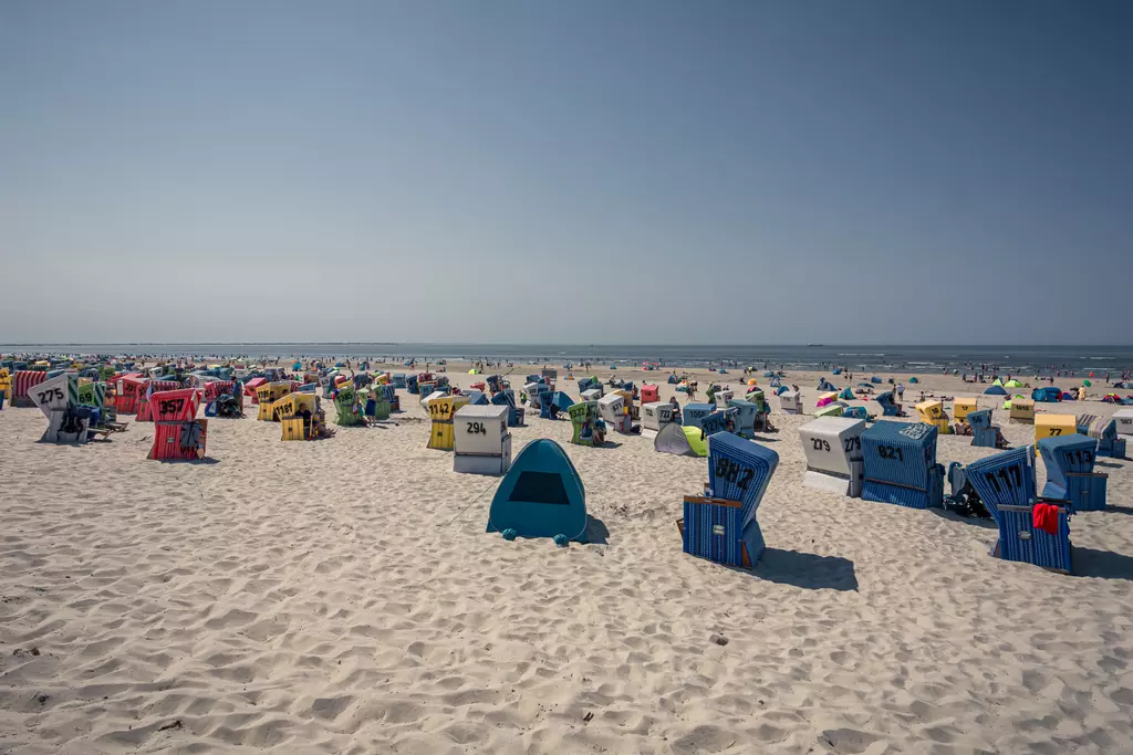 Farbenfrohe Strandkörbe stehen auf einem hellen Sandstrand, im Hintergrund ist das Meer unter einem klaren Himmel sichtbar.