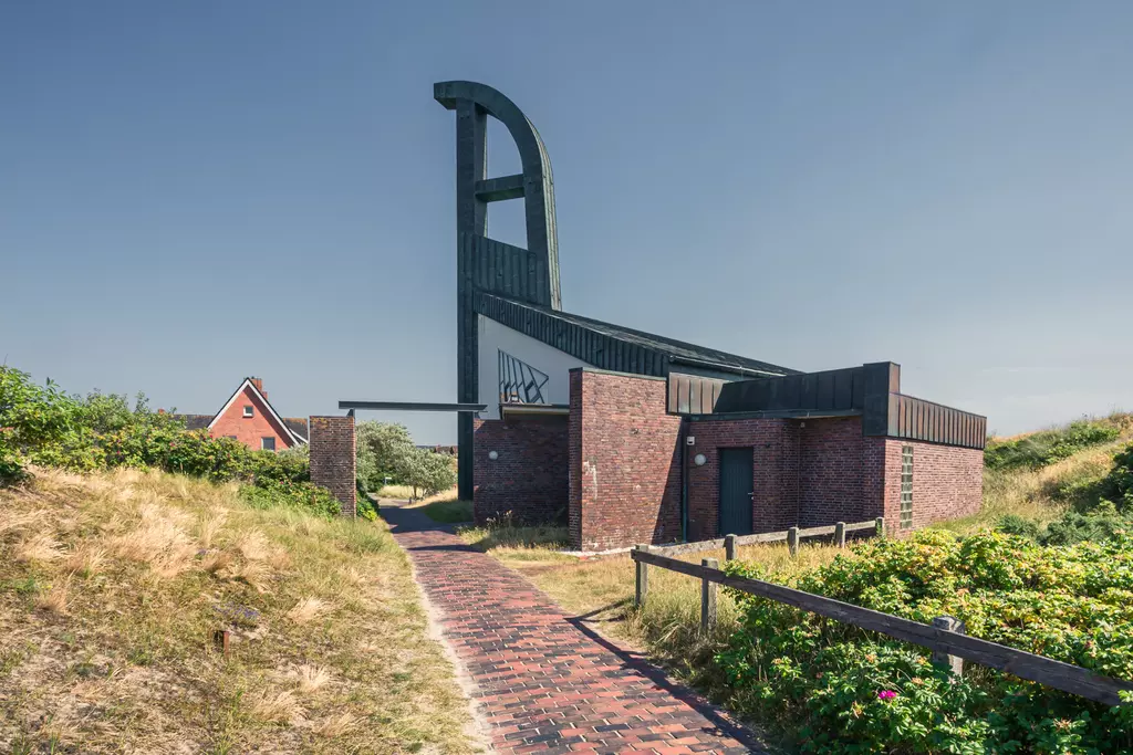 Ein moderner, architektonisch gestalteter Bau mit einer hohen, geschwungenen Wand über einem gepflasterten Weg, umgeben von Grün.