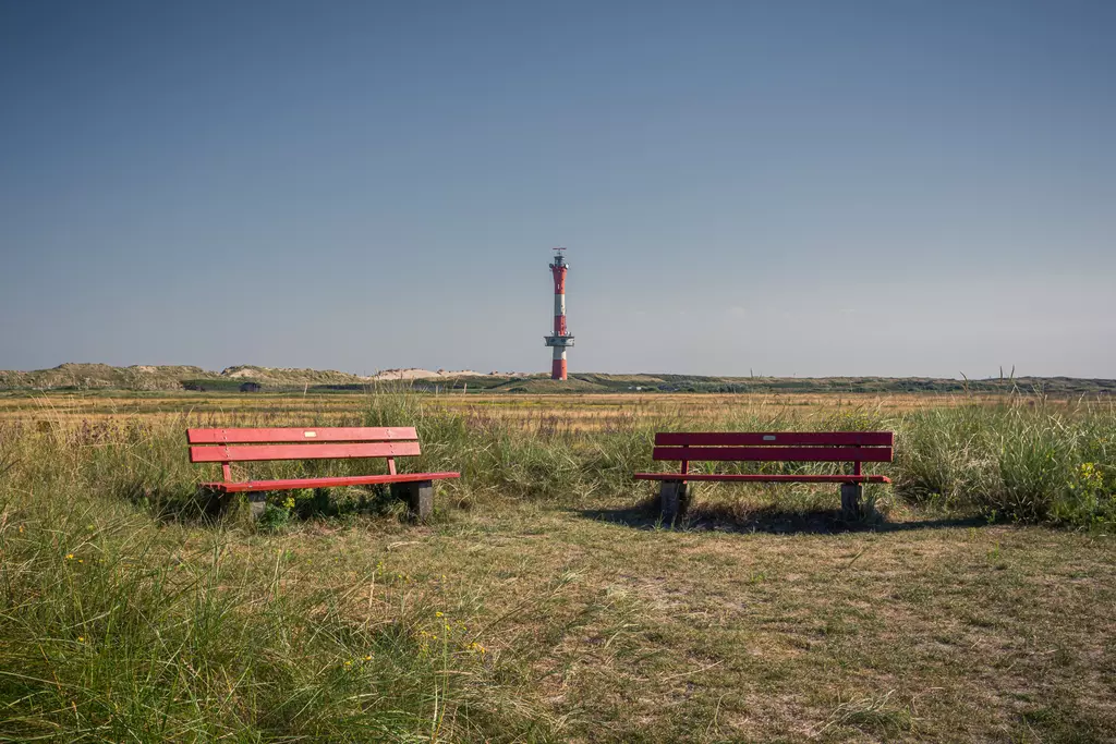 Zwei rote Bänke stehen auf einem offenen Feld, während im Hintergrund ein hoher, orange-weißer Leuchtturm sichtbar ist.