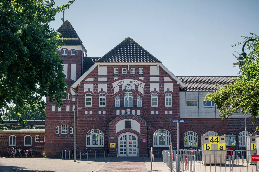 Rot-weiße Ziegelgebäudefassade mit Türmchen und vielen Fenstern, umgeben von Bäumen und Parkplatzdetails.