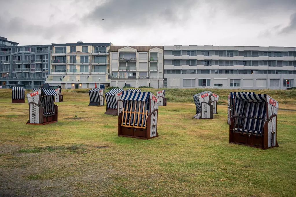 Strandkörbe stehen auf einer grünen Wiese, im Hintergrund sind moderne Gebäude unter einem bewölkten Himmel zu sehen.