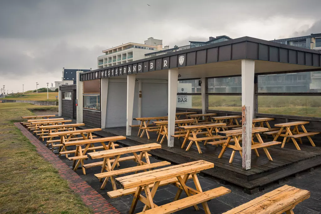 Holzterrasse mit mehreren Tischen und Bänken unter einem überdachten Bereich, umgeben von grünem Gras und einem bewölkten Himmel.