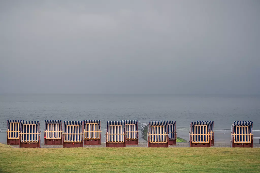 Reihen von Strandkörben stehen auf einer Wiese am Meer, die grauen Wolken reflektieren die ruhige Wasseroberfläche.