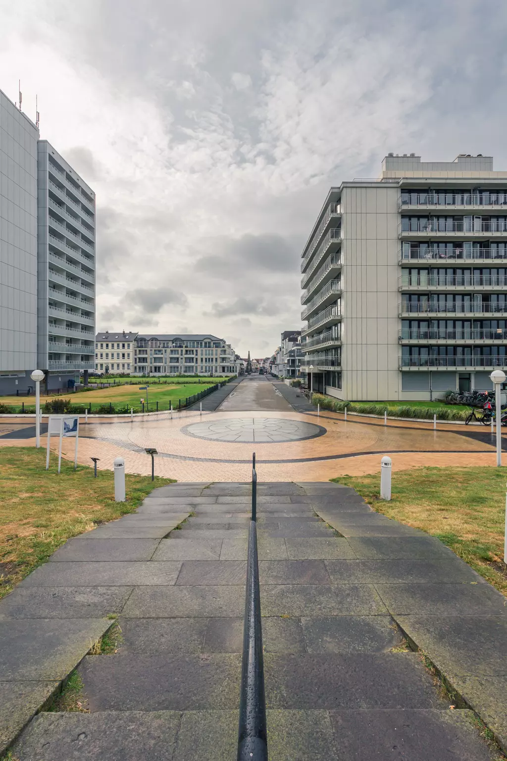 Grauer Himmel über einem breiten Weg, umgeben von modernen Gebäuden. Runder Platz in der Mitte, gesäumt von Rasenflächen.