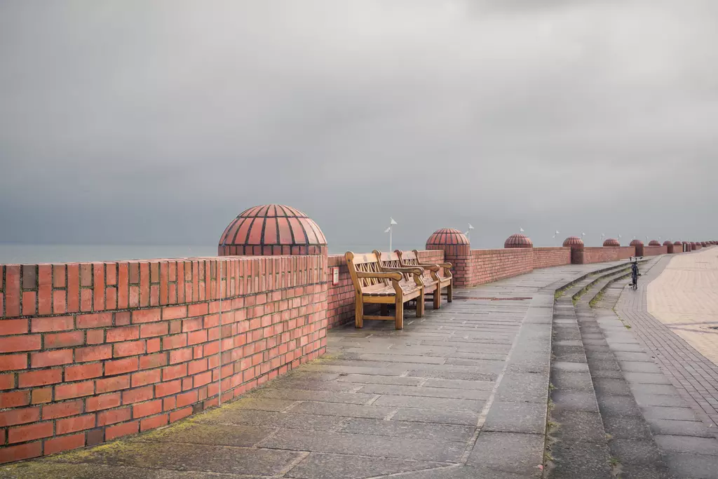 Bänke entlang einer Mauer aus roten Ziegelsteinen, die in der Ferne von einem grauen Himmel und sanften Wellen umgeben sind.