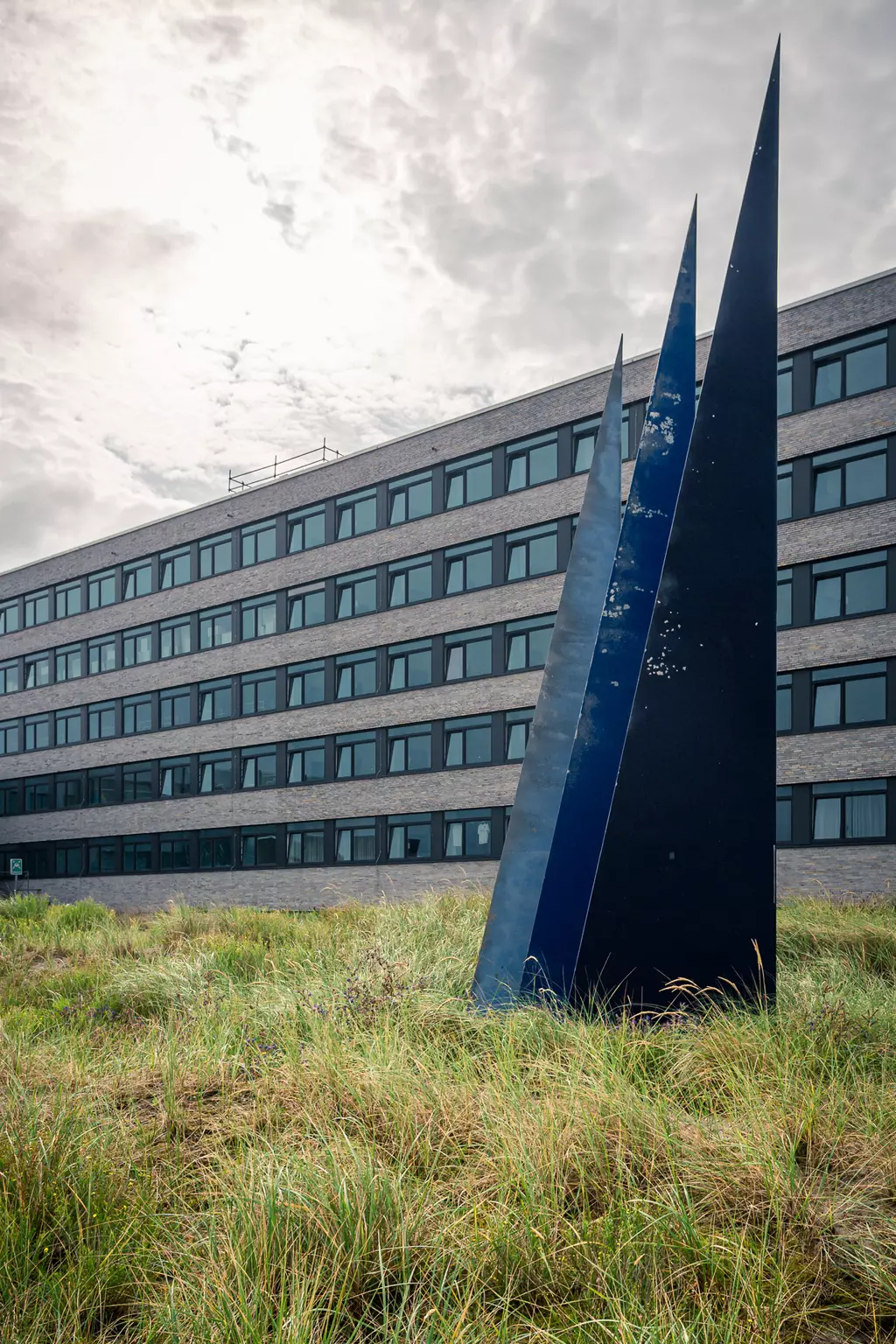 Drei spitze, schwarze Skulpturen stehen in grünem Gras vor einem modernen Gebäude mit vielen Fenstern. Der Himmel ist bewölkt.