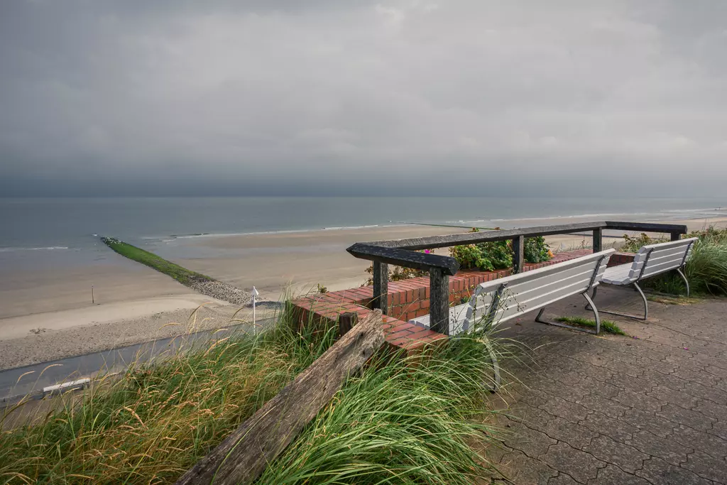 Eine Aussichtskanzel mit Bänken über einem ruhigen Strand, grauer Himmel und sanfte Wellen im Hintergrund, umgeben von hohem Gras.