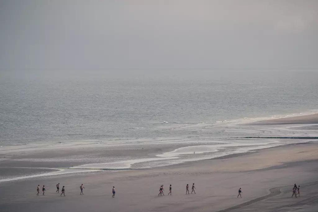 Mehrere Menschen spazieren am Strand, während sanfte Wellen den Meereshorizont umspülen und der Himmel bewölkt ist.