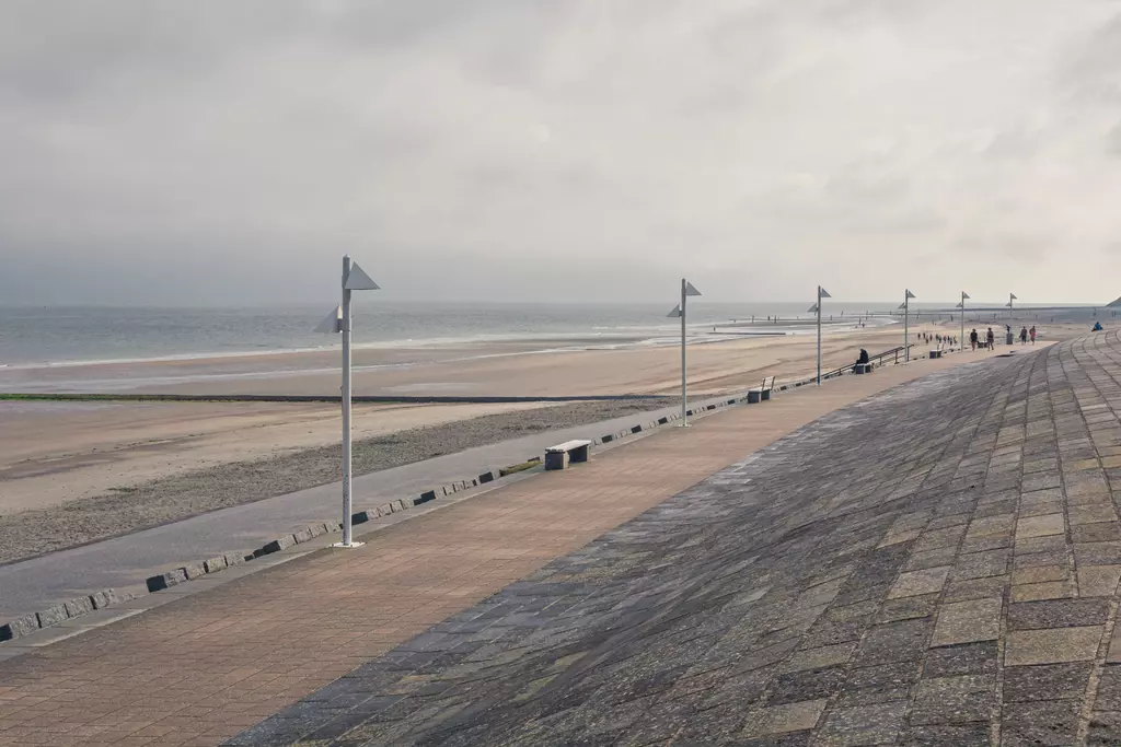 Ein breiter Strandpromenade mit grauem Himmel, Windfahnen und wenigen Menschen am Horizont entlang des strahlend leeren Strands.