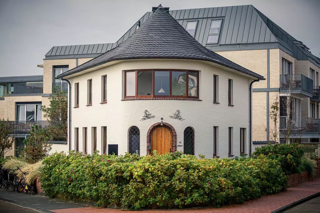 Rundes weißen Haus mit einem schwarzen Dach, umgeben von grünen Sträuchern und modernen Gebäuden im Hintergrund.