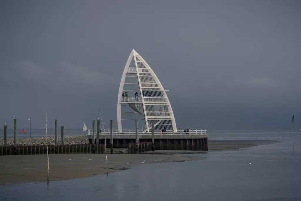 Ein moderner, weiße Aussichtsplattform in Form eines Segels befindet sich an der Küste, umgeben von ruhigem Wasser und grauem Himmel.