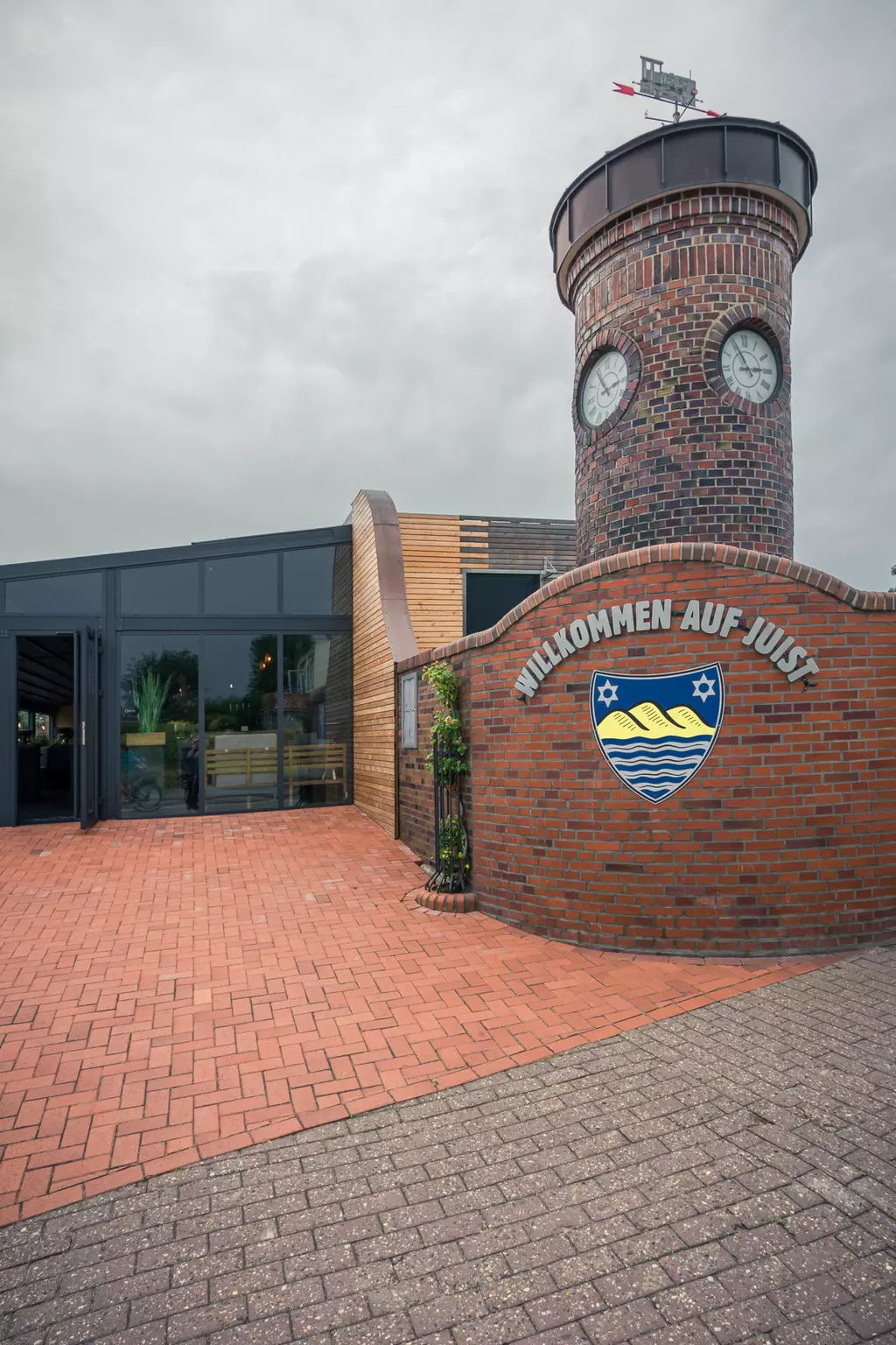 Hinter einer Backsteinmauer steht ein Turm mit zwei Uhren und einem Schild, das „Willkommen auf Juist“ zeigt, unter einem grauen Himmel.