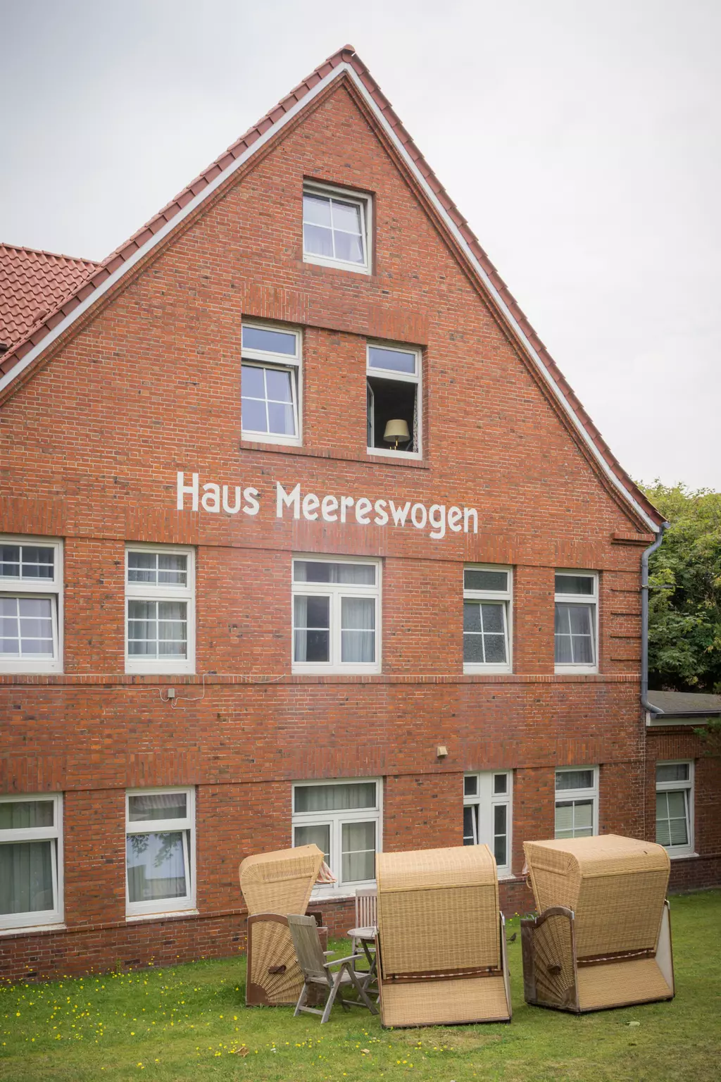 Eine zweigeschossige Backsteinfassade mit der Aufschrift Haus Meereswogen. Vor dem Gebäude stehen zwei Strandkörbe.