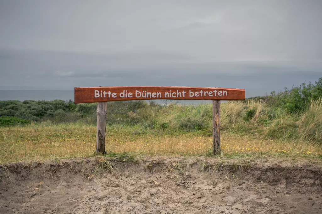 Ein Holzschild mit der Aufschrift Bitte die Dünen nicht betreten steht im Vordergrund, umgeben von Gras und einem Küstenhorizont.