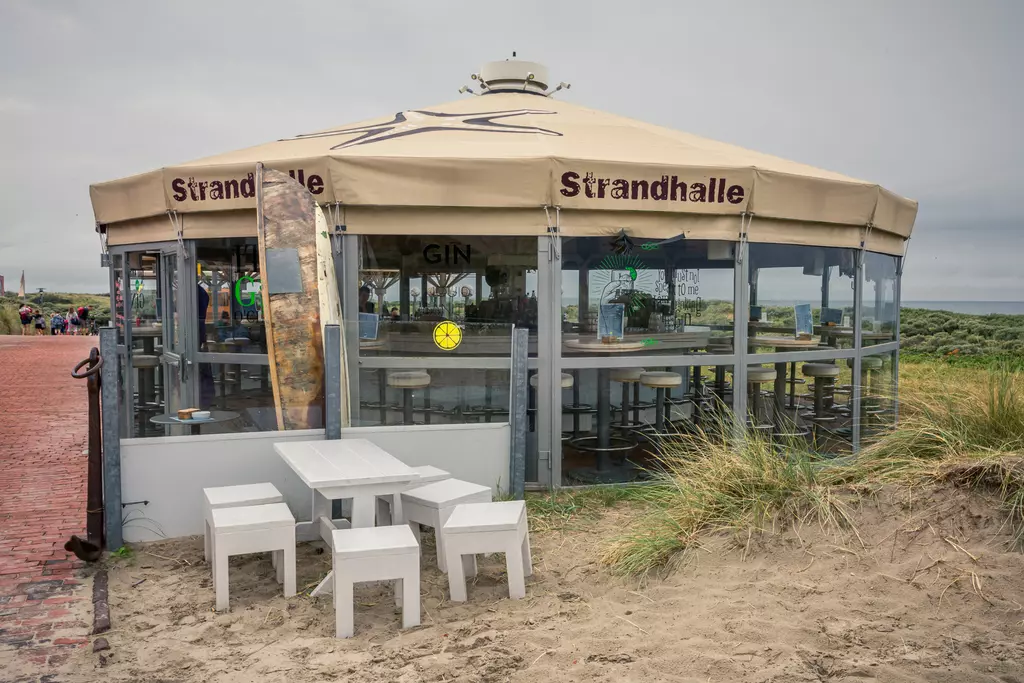 Ein rundes Strandcafé mit großem Sonnendach steht am Küstensand, umgeben von Tischen und Stühlen.