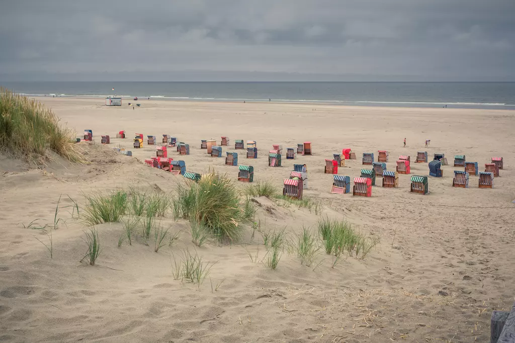 Bunte Strandkörbe stehen auf einem weitläufigen Sandstrand, umgeben von sanften Dünen und einem bewölkten Himmel.