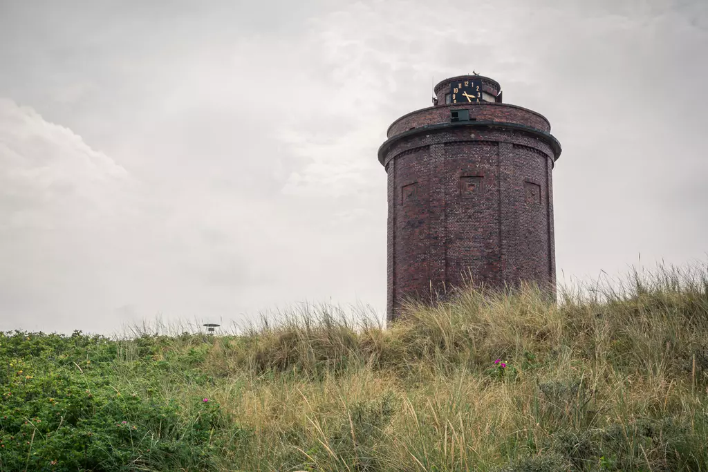 Ein roter Ziegelbau mit einer Uhr auf dem Dach steht auf einer Wiese, umgeben von grünem Grass und unter einem bewölkten Himmel.