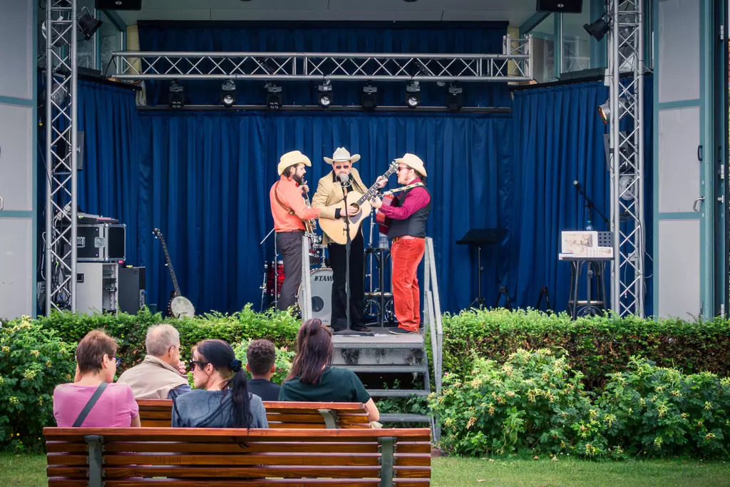 Bühne mit drei Musikern in Cowboy-Outfits, die ein Konzert geben. Das Publikum sitzt auf einer Bank im Vordergrund.