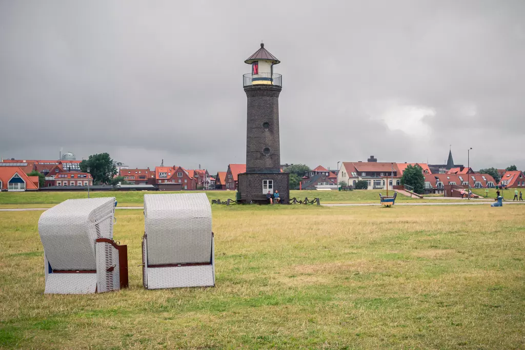 Ein Leuchtturm steht auf einer Wiese, während zwei Strandkörbe davor platziert sind. Im Hintergrund sind Häuser und ein bewölkter Himmel sichtbar.