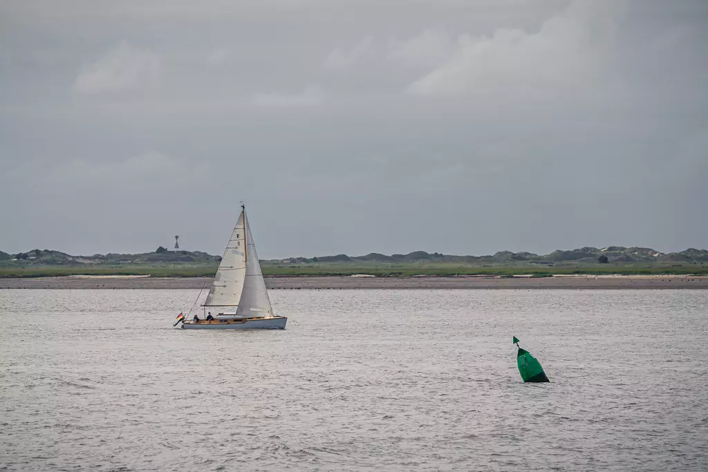 A sailboat glides over calm water near a green navigation buoy, under a cloudy sky.