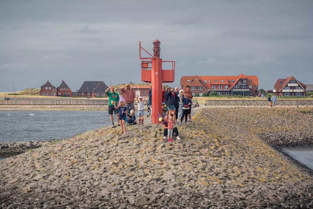 A group of people stands on a gravel-covered jetty near a red navigation buoy, with a beach visible in the background.