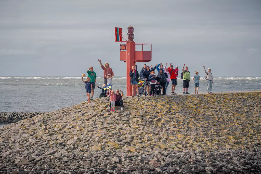 A group of people stands on a stone jetty, waving at the camera, with a red lighthouse signal and the sea behind them.