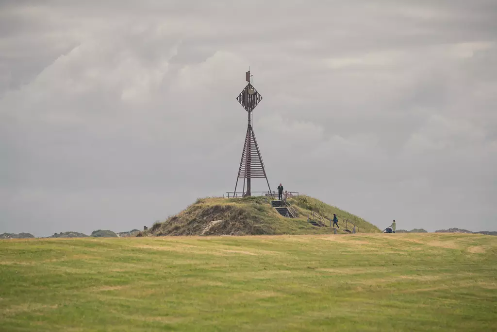 A small hill with a tall metal signal mast on top, surrounded by green grass and a cloudy sky.