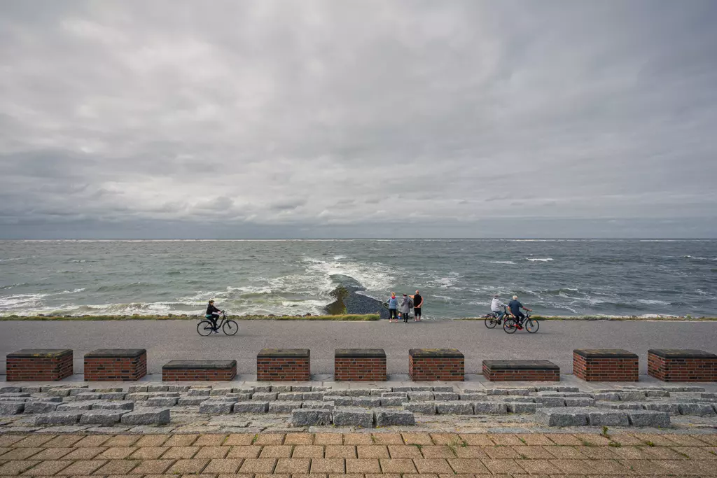 A promenade with cyclists, a group of people by the sea, and gray clouds in the sky.