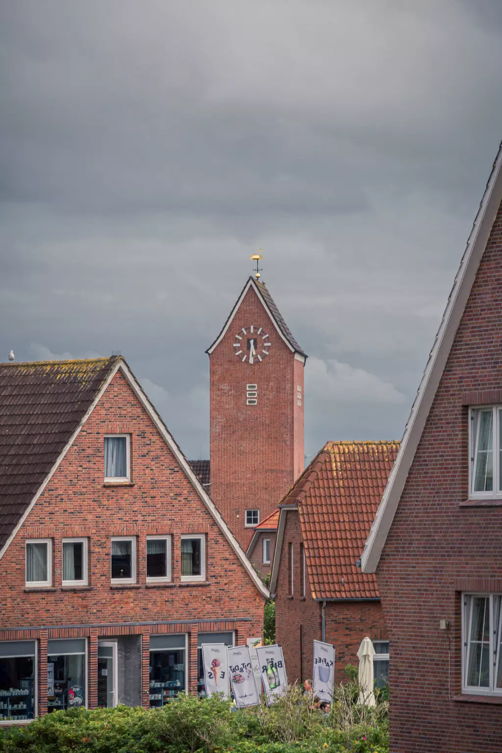 Red brick houses with gabled roofs and a church tower featuring a clock and weathervane under a cloudy sky.