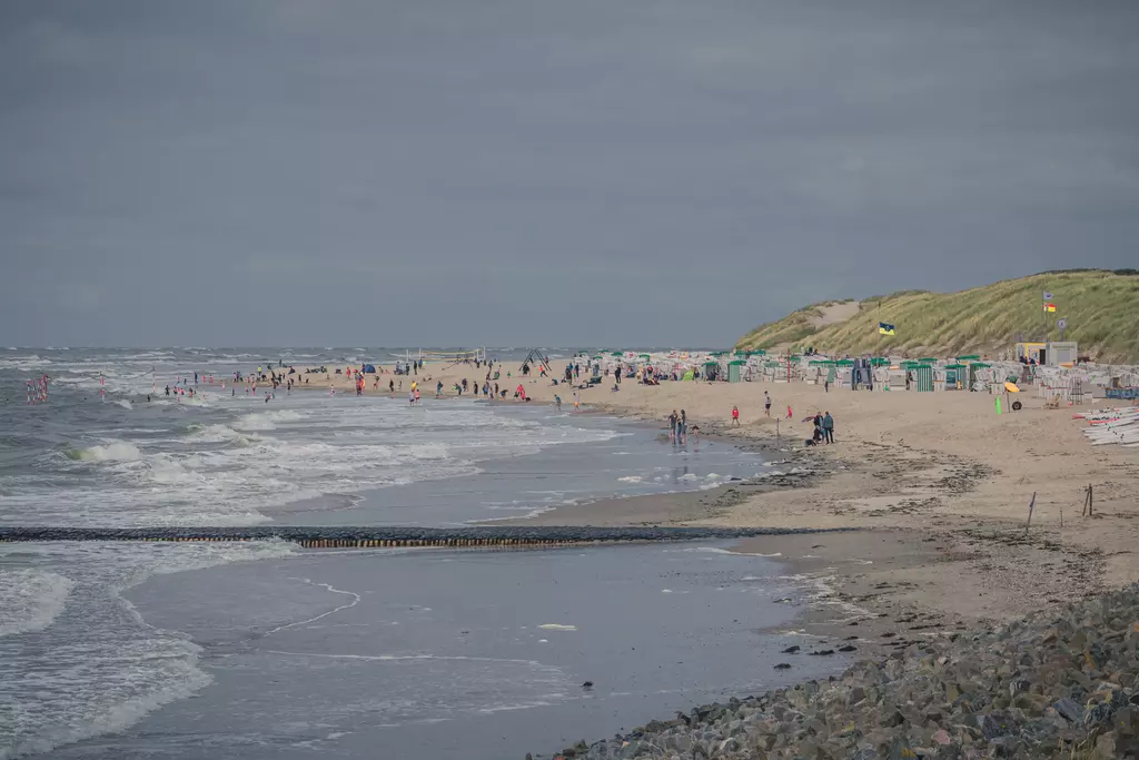 People walk along the beach as waves hit the sandy shore. Colorful beach cabins line up in the background.