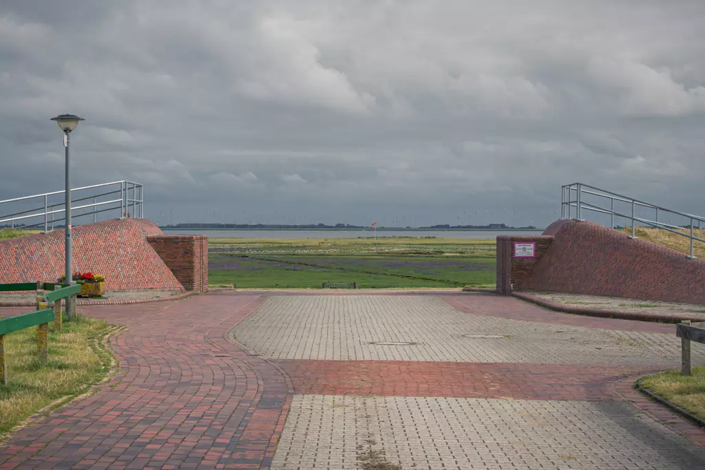 Two brick ramps lead from a paved path to a wide, open landscape featuring sky and water.