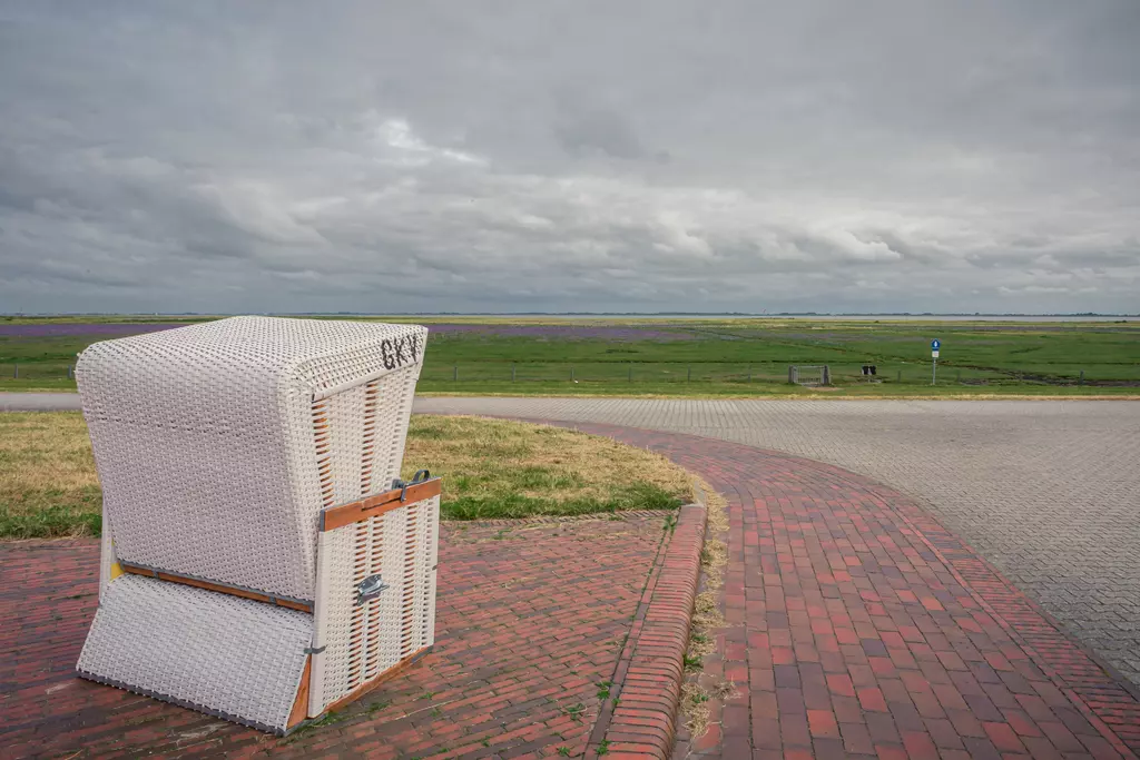 A white beach chair sits on a paved path, surrounded by green grass and a gray sky filled with clouds.