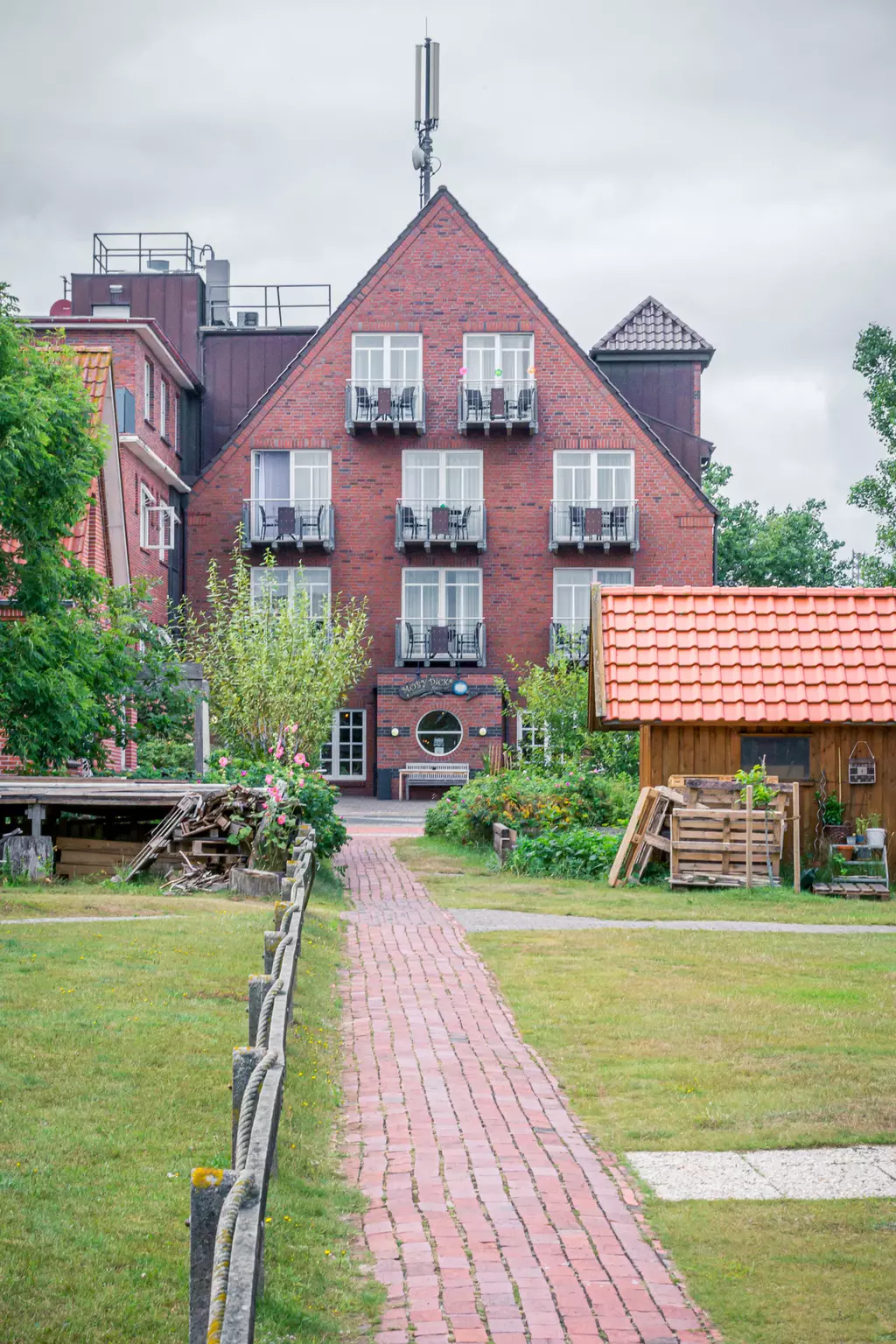 A brick walkway leads to a red brick building with balconies. Trees and a wooden structure are in the foreground.