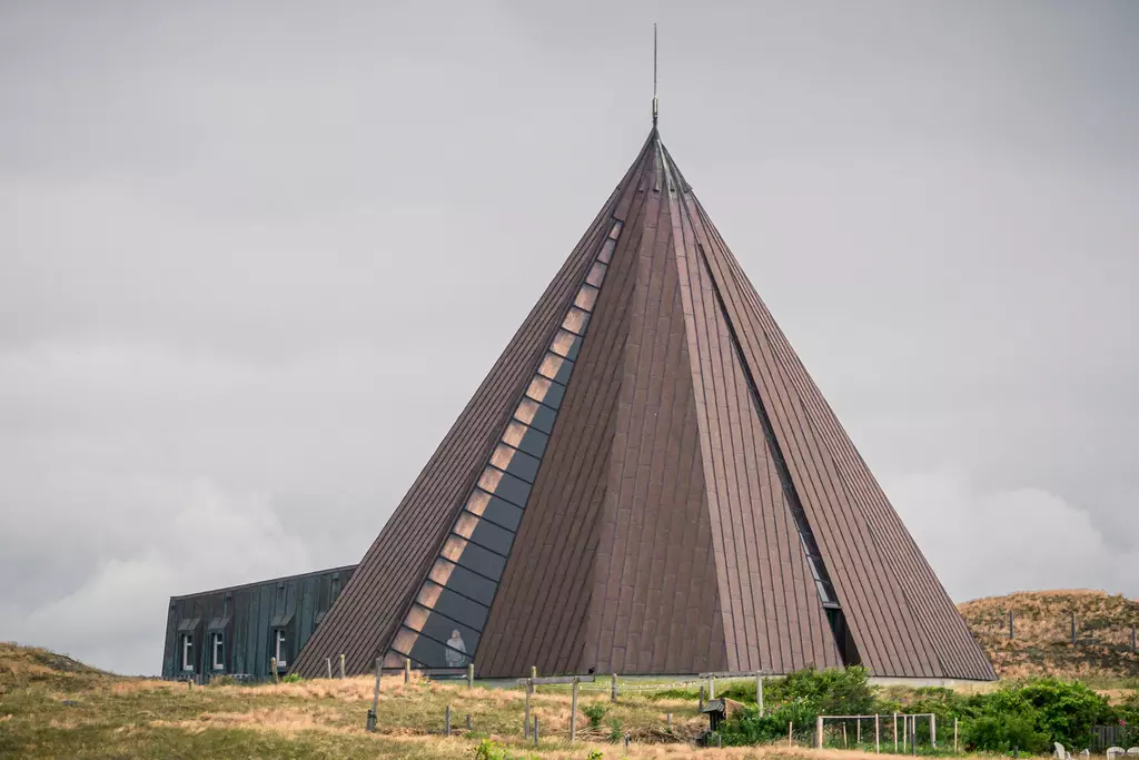 A striking pyramid-shaped building with sloped walls and a pointed top, surrounded by gentle hills and greenery.