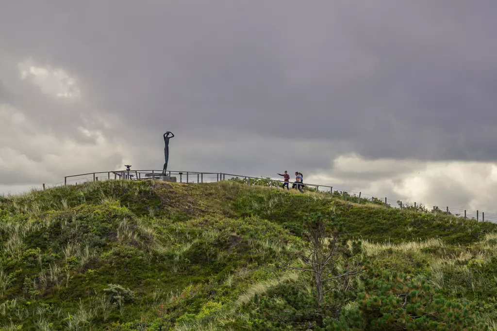 A green hillside with a statue on a plateau and two people nearby, under a cloudy sky.