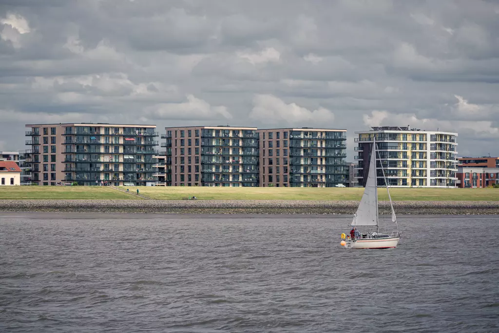 Moderne Wohngebäude am Ufer mit Rasenfläche und Segelboot im Vordergrund auf ruhigem Wasser unter bewölbtem Himmel.