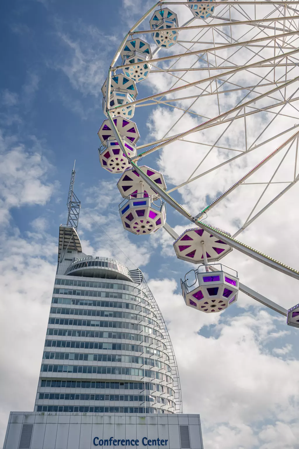 Ein großes Riesenrad mit bunten Kabinen neben einem modernen Gebäude mit glatter Fassade und dem Schriftzug "Conference Center".