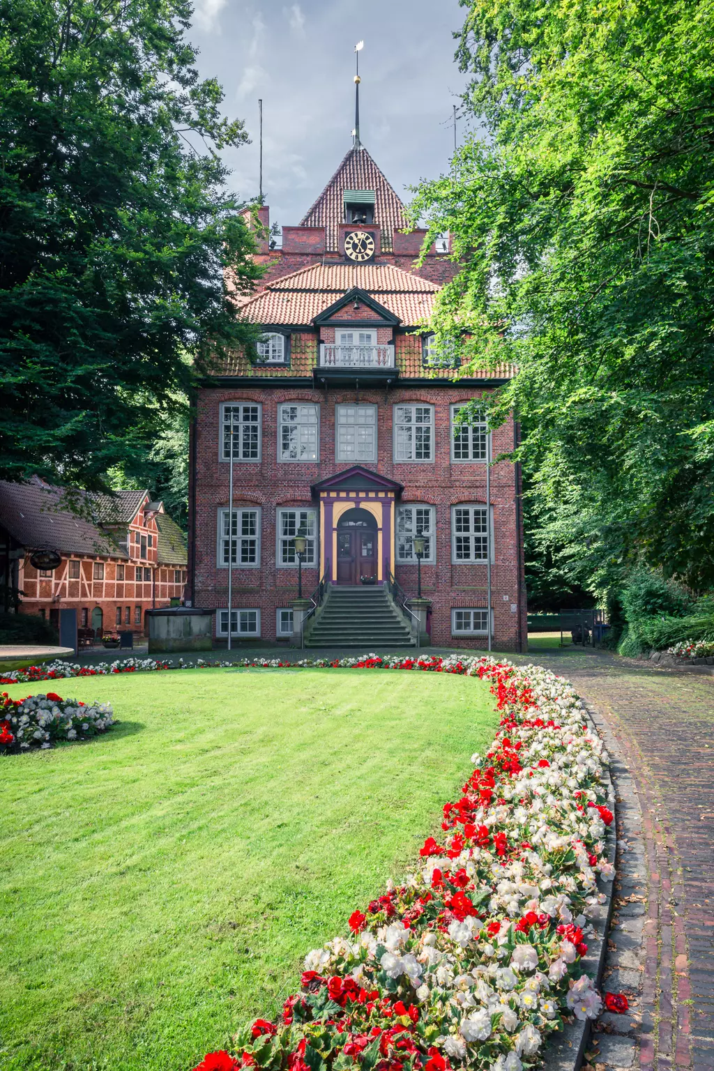 Ein imposantes, dreigeschossiges, rotes Backsteingebäude mit einem spitzen Dach und einem Eingang mit Treppe vor einem gepflegten Garten.