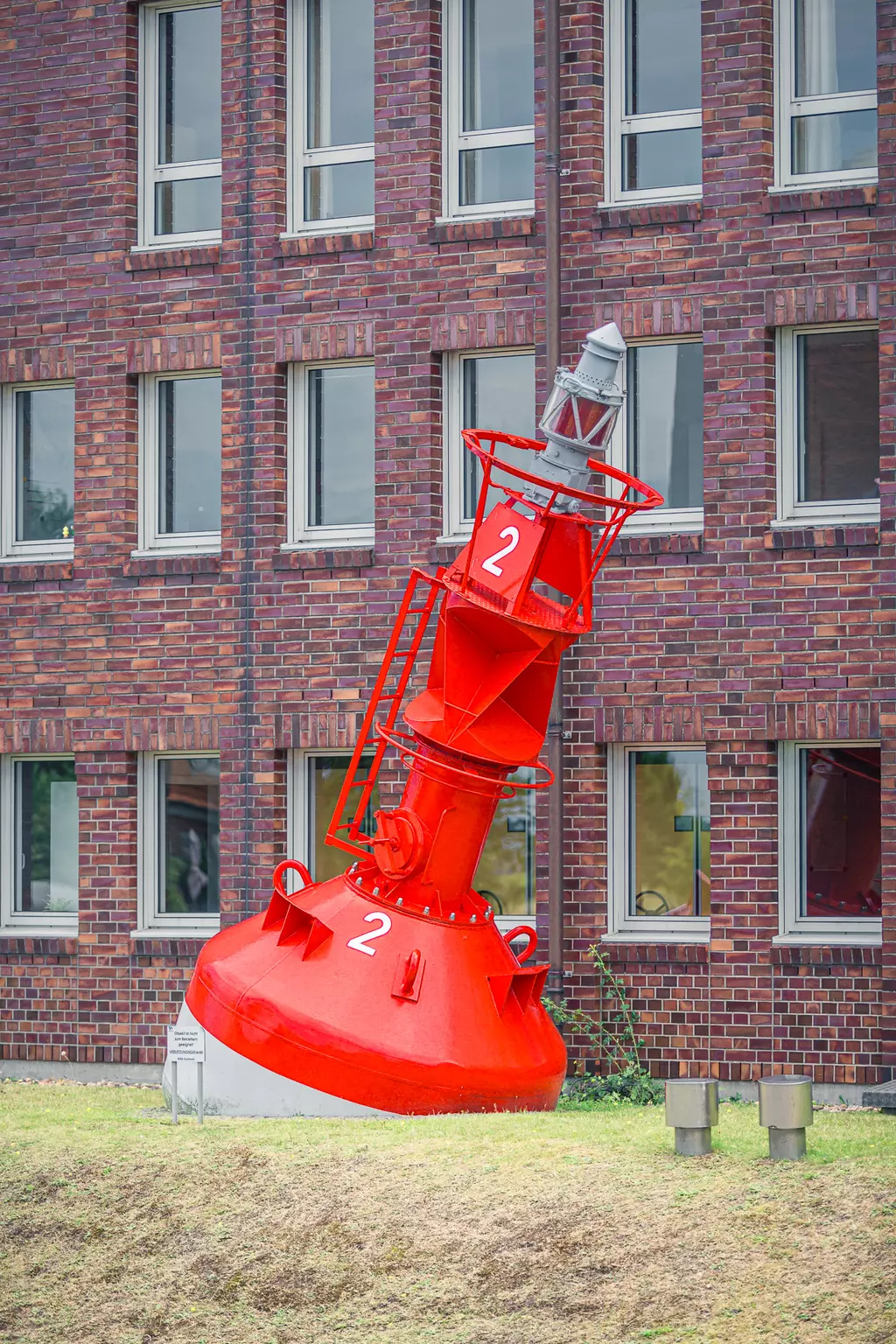 Ein roter Hafenfeuerkörper in schräger Lage vor einem Backsteingebäude mit mehreren Fenstern.