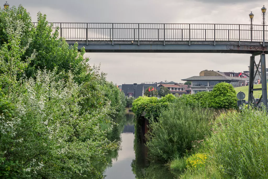 A metal bridge crosses a narrow canal, surrounded by lush greenery and buildings in the background.