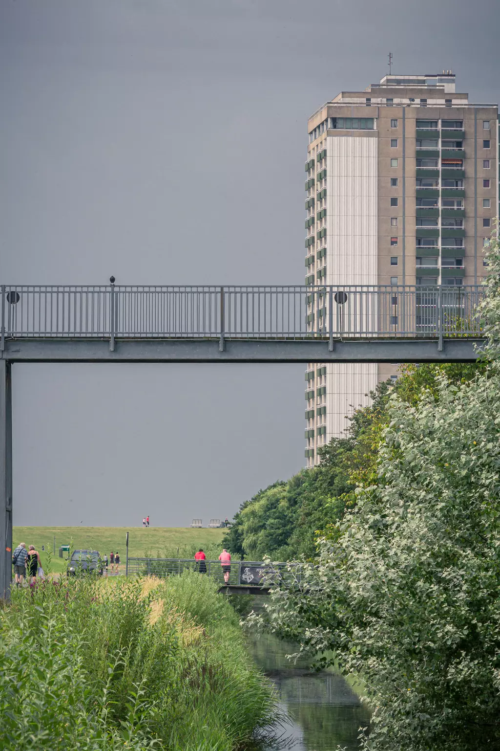 A tall residential building stands next to a footbridge over a narrow canal, with green banks and trees in the background.